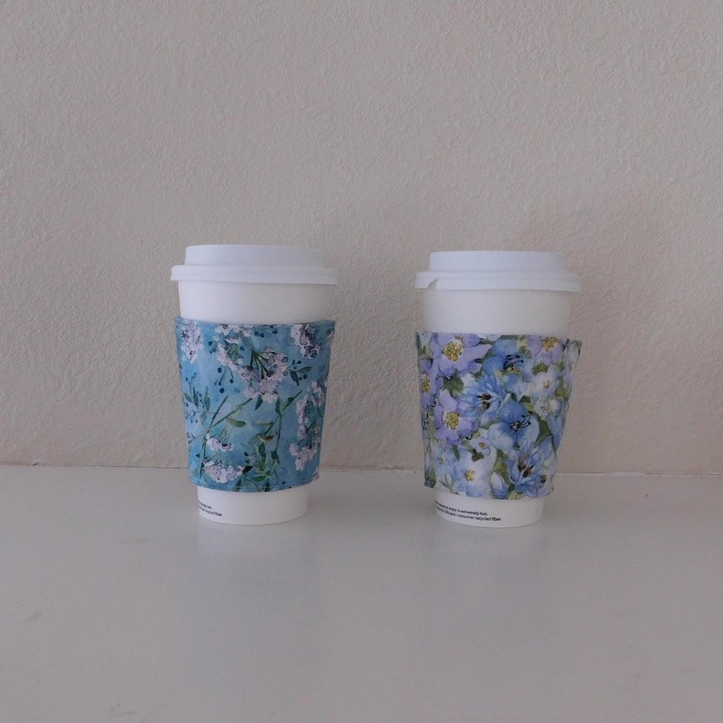 May include: Two white disposable coffee cups with lids, each featuring a fabric sleeve. The left sleeve has a light blue background with small white and pink floral patterns. The right sleeve displays a dense pattern of blue and purple flowers.