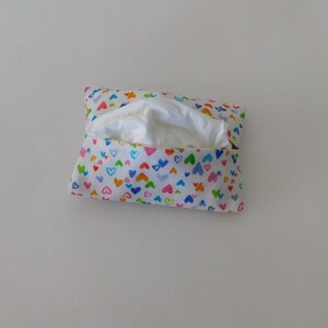 May include: A rectangular fabric tissue holder featuring a white background with a repeating pattern of small, hand-drawn hearts in various colours including blue, green, orange, pink, and yellow, with a white tissue visible in the centre opening.