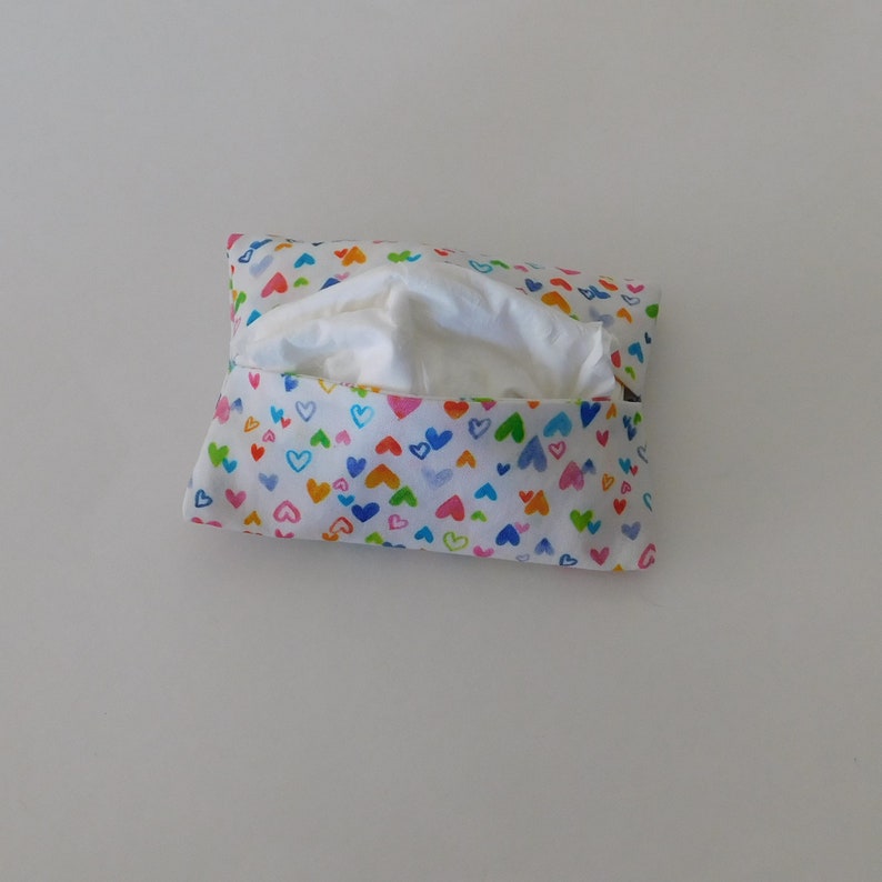 May include: A rectangular fabric tissue holder featuring a white background with a repeating pattern of small, hand-drawn hearts in various colours including blue, green, orange, pink, and yellow, with a white tissue visible in the centre opening.