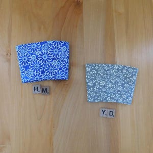 May include: Two fabric cup sleeves. One sleeve features a bright blue background with a white daisy floral pattern. The second sleeve has a gray background with a white intricate floral vine pattern. Each sleeve is paired with two Scrabble tiles.