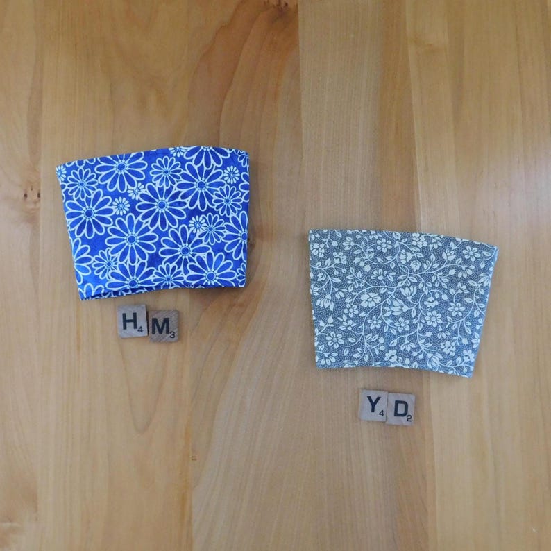 May include: Two fabric cup sleeves. One sleeve features a bright blue background with a white daisy floral pattern. The second sleeve has a gray background with a white intricate floral vine pattern. Each sleeve is paired with two Scrabble tiles.