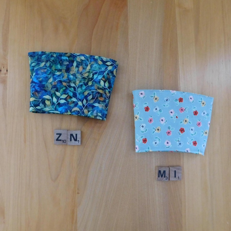 May include: Two fabric cup sleeves. The left sleeve features a dark blue and green leafy botanical pattern with a wooden tile labeled ZN. The right sleeve is light blue with a small red and yellow floral print and a wooden tile labeled MI.