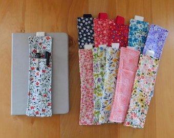 May include: A collection of rectangular fabric pen holders featuring various floral and botanical patterns in colours including blue, red, pink, yellow, and purple. Each holder has a small elastic loop at the top and one is attached to a grey notebook.