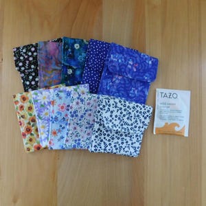 May include: A collection of ten small fabric pouches featuring various floral and patterned designs in colors including blue, purple, yellow, and black, arranged in two rows next to a white Tazo Wild Sweet Orange tea bag packet.