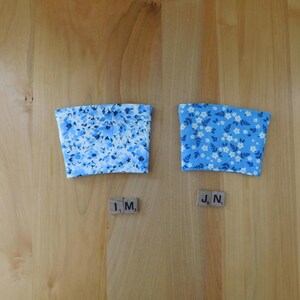 May include: Two fabric cup sleeves with a tapered shape. The left sleeve features a light blue floral pattern with darker blue accents. The right sleeve has a bright blue background with a repeating pattern of small white flowers and green leaves.