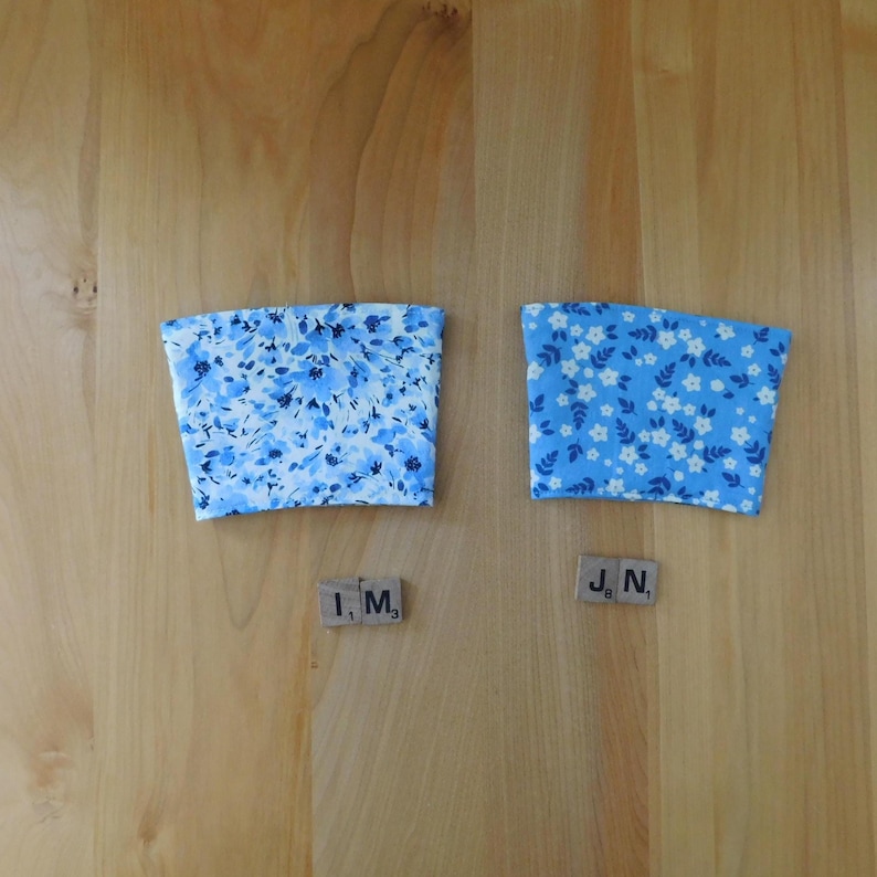 May include: Two fabric cup sleeves with a tapered shape. The left sleeve features a light blue floral pattern with darker blue accents. The right sleeve has a bright blue background with a repeating pattern of small white flowers and green leaves.