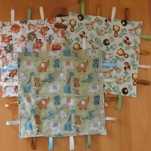 May include: Four baby security blankets with animal patterns. One has elephants, giraffes, and crocodiles on a green background. Others feature lions, bears, and foxes. Each blanket has colorful ribbons attached to the edges.