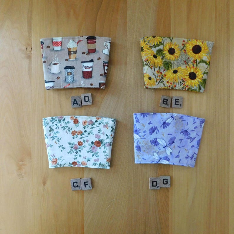 May include: Four fabric cup sleeves arranged in a grid. Top left features a coffee-themed print on a beige background. Top right displays yellow sunflowers on a tan background. Bottom left shows a floral pattern. Bottom right features purple flowers.