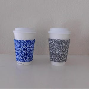 May include: Two white disposable coffee cups with lids, each featuring a fabric sleeve. One sleeve is bright blue with a white daisy pattern, and the other sleeve is dark gray with a dense, intricate white floral and vine print design.