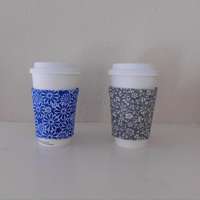 May include: Two white disposable coffee cups with lids, each featuring a fabric sleeve. One sleeve is bright blue with a white daisy pattern, and the other sleeve is dark gray with a dense, intricate white floral and vine print design.