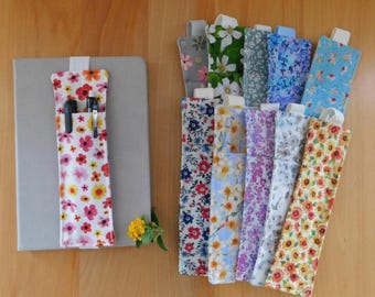 May include: A collection of rectangular fabric pen holders featuring various floral patterns in colours including pink, yellow, blue, and green. One holder is attached to a grey notebook with two black pens inserted, while others are stacked nearby.