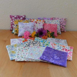 May include: A collection of small, square fabric sachets featuring various floral and botanical patterns in shades of purple, pink, yellow, blue, and white. The items are arranged in a layered pile, with some standing upright and others lying flat.