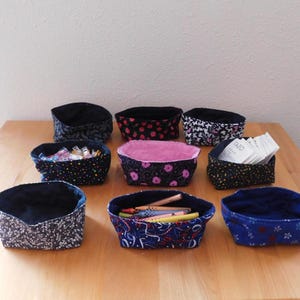 May include: Nine fabric storage baskets in various patterns and colors, including floral, polka dot, and paisley designs. Some baskets contain items like pens, sewing clips, and tea bags. The baskets are arranged on a wooden table.