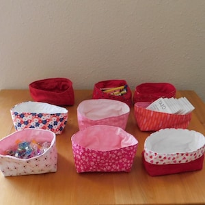 May include: A collection of nine fabric storage baskets in various colors and patterns. The baskets are red, pink, and patterned with floral and heart designs. Some baskets contain small items like tea bags and sewing supplies. They are arranged on a wooden surface.