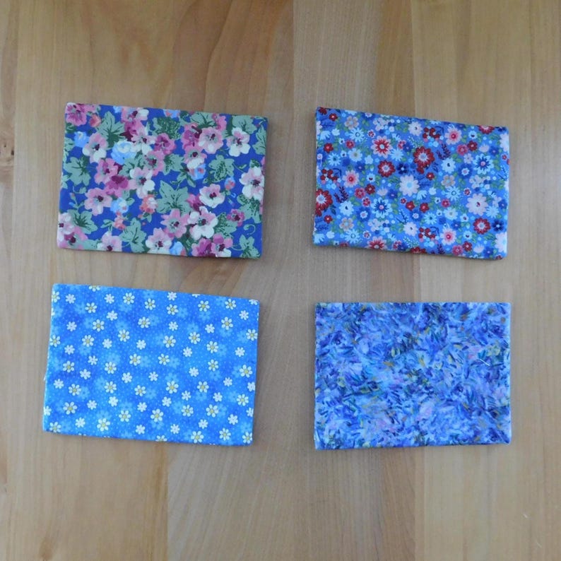 May include: Four rectangular fabric coasters featuring distinct blue-toned floral patterns. The top left has pink and white flowers on blue, the top right has small red and blue blossoms, the bottom left has white daisies, and the bottom right has blue petals.
