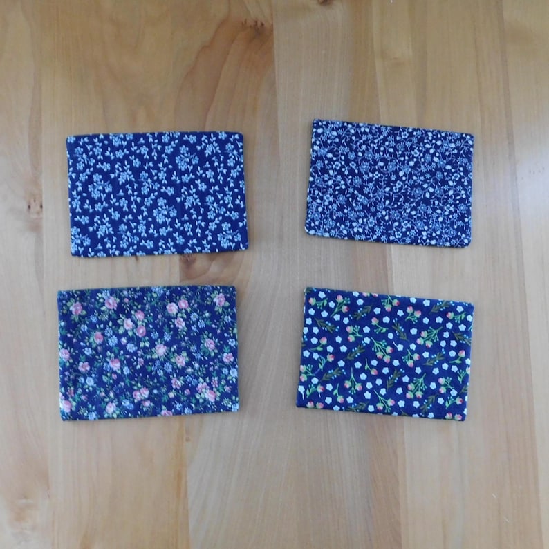 May include: A set of four rectangular fabric coasters featuring dark blue backgrounds with distinct floral patterns. Two coasters display small light blue blossoms, one features pink and blue flowers, and one shows white and green floral motifs.