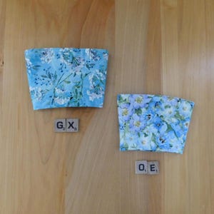 May include: Two fabric cup sleeves featuring a light blue floral pattern with white and dark blue blossoms and green foliage. The left sleeve is accompanied by two wooden tiles marked G2 and X8, while the right sleeve has tiles marked O1 and E1.