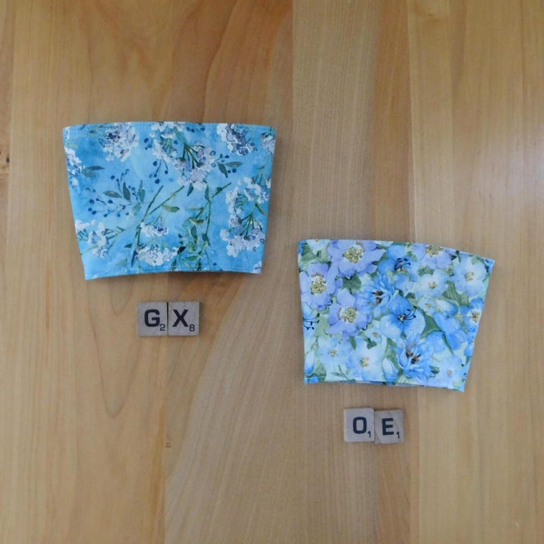 May include: Two fabric cup sleeves featuring a light blue floral pattern with white and dark blue blossoms and green foliage. The left sleeve is accompanied by two wooden tiles marked G2 and X8, while the right sleeve has tiles marked O1 and E1.