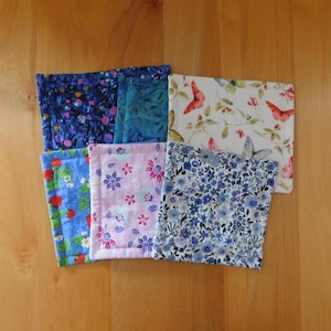 May include: A collection of seven square fabric coasters with various floral and butterfly patterns. The coasters feature a range of colors, including blue, pink, white, and red. Each coaster is approximately 4 inches square.