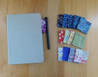May include: A light grey notebook with a black pen and a purple patterned fabric bookmark, accompanied by a collection of small, square fabric swatches featuring various floral and geometric patterns in blue, red, yellow, green, and pink.