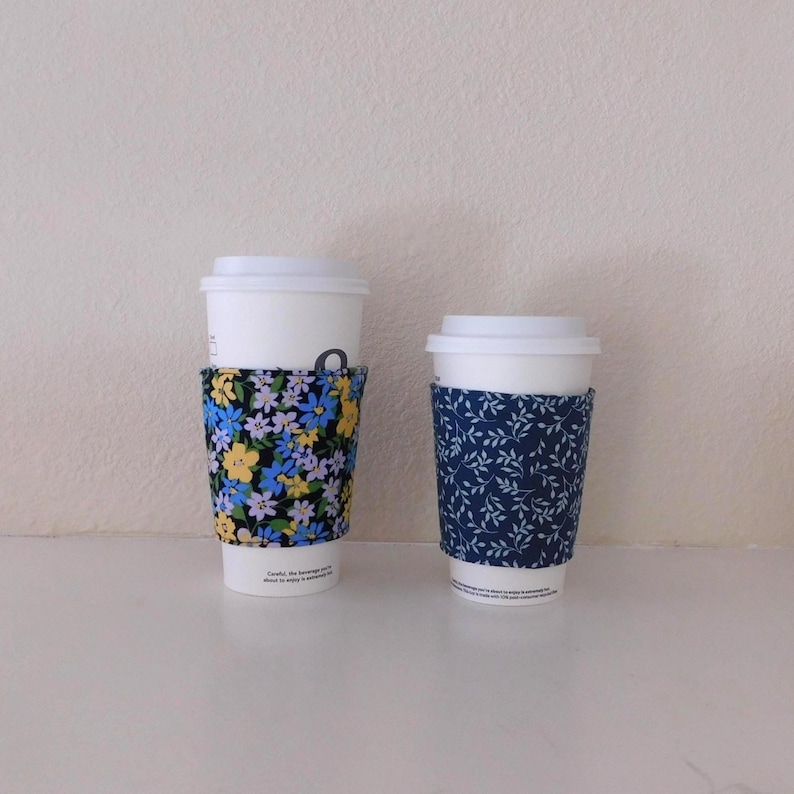 May include: Two white disposable coffee cups with lids, each featuring a fabric sleeve. The larger cup has a black sleeve with a yellow, blue, and light purple floral pattern. The smaller cup has a dark blue sleeve with a white leaf pattern.