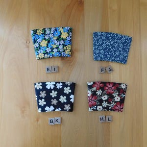 May include: Four fabric cup sleeves with distinct patterns: one with blue and yellow flowers, one with a dark blue leaf print, one with white flowers on black, and one with red and white flowers on black. Each sleeve is labeled with wooden letter tiles.