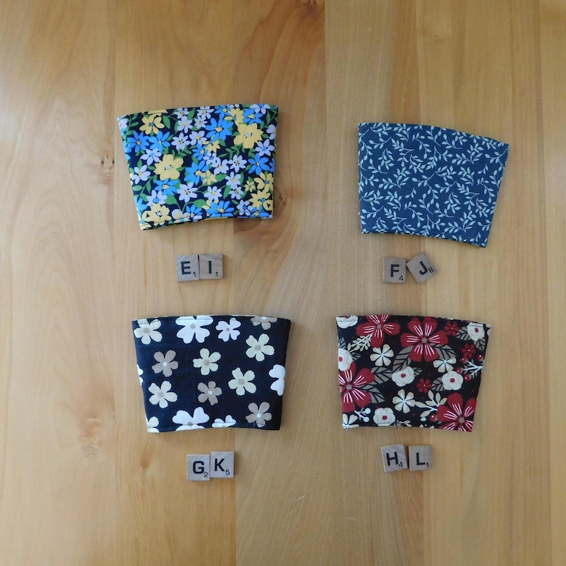 May include: Four fabric cup sleeves with distinct patterns: one with blue and yellow flowers, one with a dark blue leaf print, one with white flowers on black, and one with red and white flowers on black. Each sleeve is labeled with wooden letter tiles.