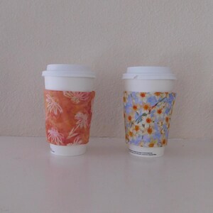 May include: Two white disposable coffee cups with lids, each featuring a fabric sleeve. The left sleeve is orange with a white floral pattern, while the right sleeve is light blue with a pattern of yellow and white daffodils and green stems.