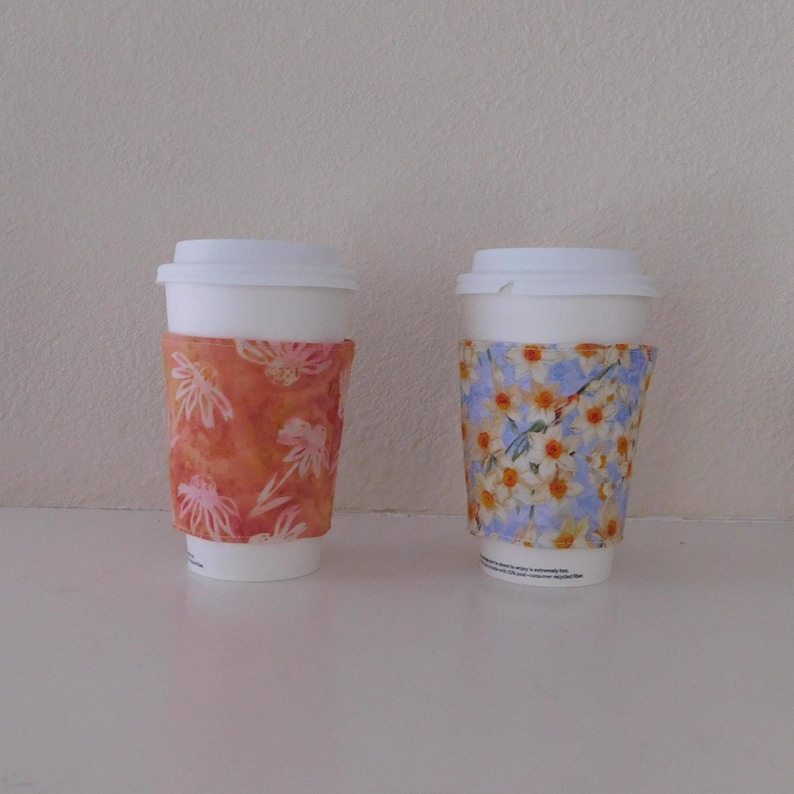 May include: Two white disposable coffee cups with lids, each featuring a fabric sleeve. The left sleeve is orange with a white floral pattern, while the right sleeve is light blue with a pattern of yellow and white daffodils and green stems.