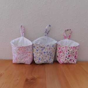 May include: Three fabric hanging baskets with white interiors and floral exterior patterns. The left basket features small pink and orange flowers, the center has a mix of blue and purple blossoms, and the right has larger pink and red floral prints.