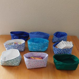 May include: A collection of nine fabric storage baskets in various colors and patterns, including blue floral, solid blue, dark blue with stars, light blue with white flowers, pink with small dots, and solid green, some containing small items.