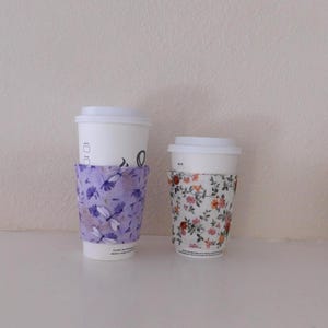 May include: Two white disposable coffee cups with lids, each featuring a fabric sleeve. The larger cup has a light purple sleeve with a dragonfly and floral pattern, while the smaller cup has a white sleeve with a colorful floral print.