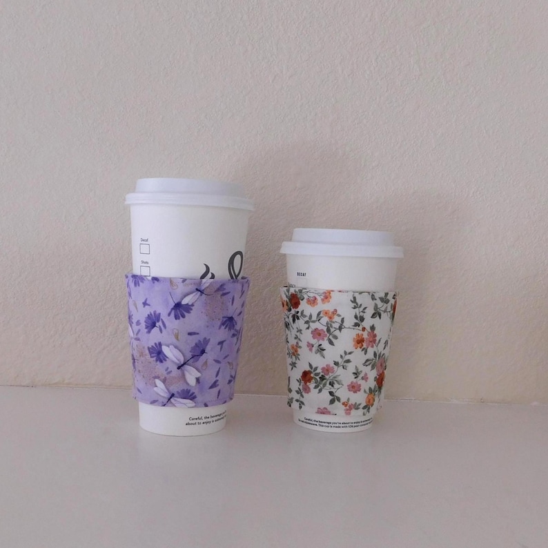 May include: Two white disposable coffee cups with lids, each featuring a fabric sleeve. The larger cup has a light purple sleeve with a dragonfly and floral pattern, while the smaller cup has a white sleeve with a colorful floral print.