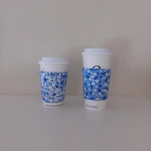 May include: Two white disposable coffee cups with lids, each featuring a fabric sleeve. The smaller cup has a blue and white floral pattern, while the taller cup features a blue sleeve with a repeating pattern of small white flowers and leaves.