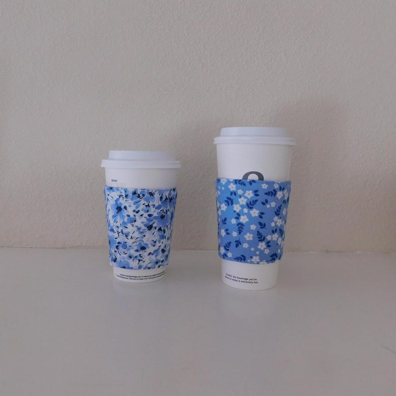May include: Two white disposable coffee cups with lids, each featuring a fabric sleeve. The smaller cup has a blue and white floral pattern, while the taller cup features a blue sleeve with a repeating pattern of small white flowers and leaves.