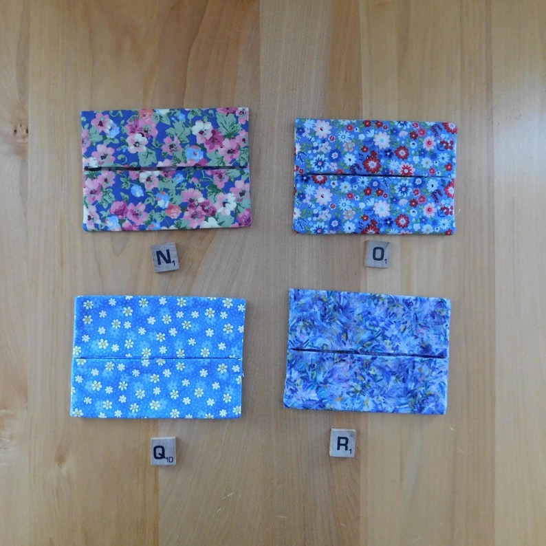 May include: Four rectangular fabric tissue holders featuring distinct blue floral patterns. Each holder has a horizontal centre opening. Small wooden tiles labelled N, O, Q, and R are placed below each corresponding fabric pouch on the surface.