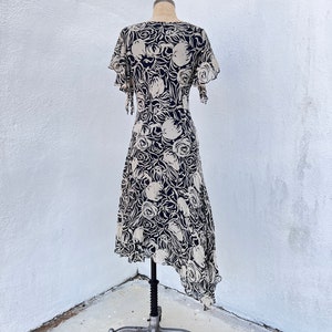 Pu&ograve; includere: Abito vintage su manichino. L'abito presenta un motivo floreale color crema su sfondo blu navy. Ha maniche corte a balze e orlo asimmetrico. L'abito &egrave; esposto su un manichino in metallo vintage.