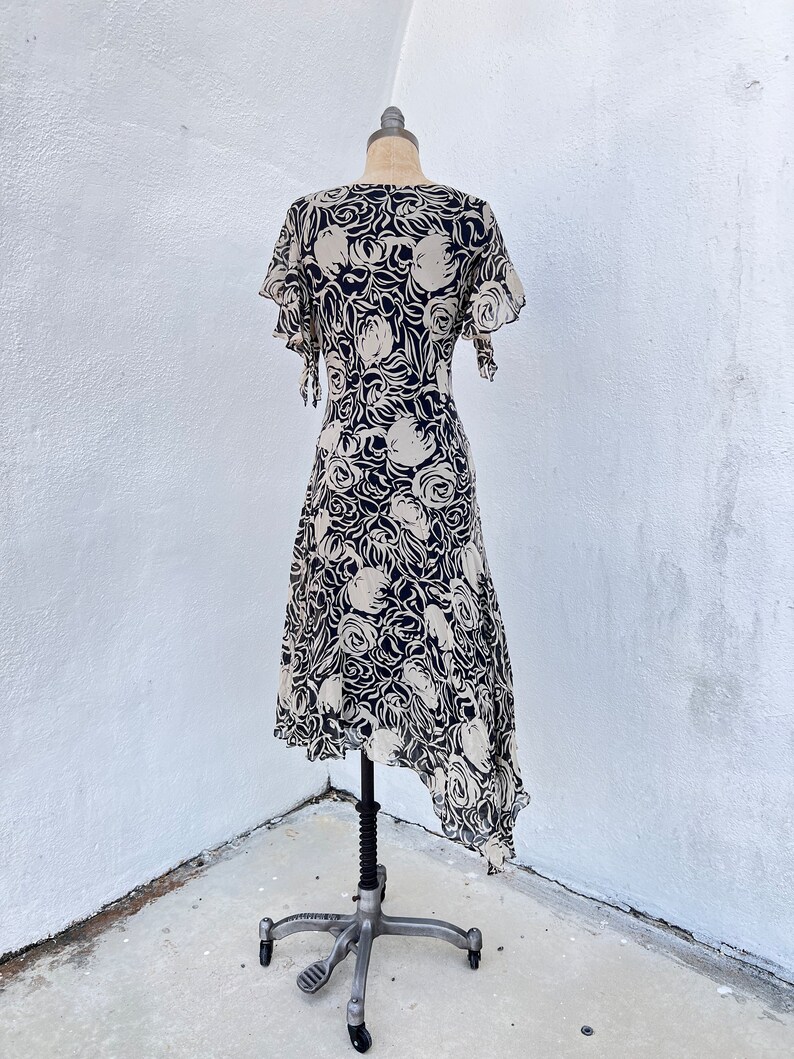 Pu&ograve; includere: Abito vintage su manichino. L'abito presenta un motivo floreale color crema su sfondo blu navy. Ha maniche corte a balze e orlo asimmetrico. L'abito &egrave; esposto su un manichino in metallo vintage.