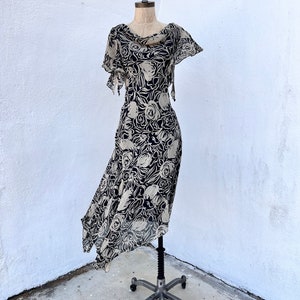 Pu&ograve; includere: Abito con stampa floreale nera e beige su manichino. L'abito ha uno scollo a cascata e un orlo asimmetrico. Il tessuto leggero presenta un motivo di rose stilizzate. L'abito &egrave; esposto contro un muro bianco.