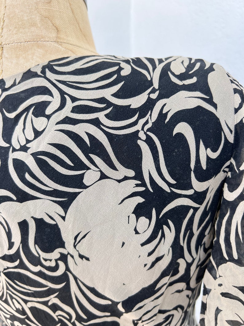 Pu&ograve; includere: Primo piano di un tessuto fantasia nero e crema. Il design presenta forme organiche a vortice in crema su sfondo nero. Il tessuto &egrave; drappeggiato su un manichino, mostrando il flusso del motivo.