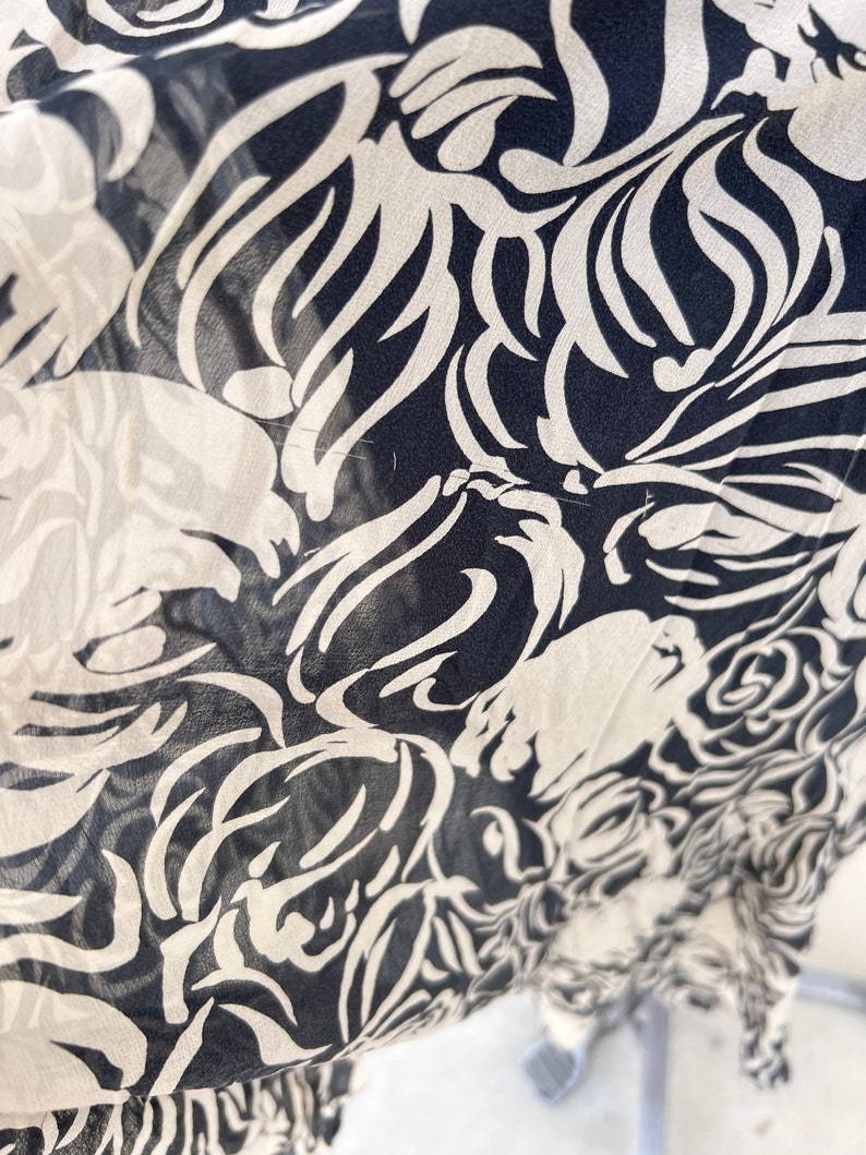 Pu&ograve; includere: Primo piano di un tessuto con un motivo floreale ripetuto. Il design presenta fiori e forme di foglie bianche stilizzate su uno sfondo blu navy scuro. Il tessuto sembra leggero e semitrasparente.