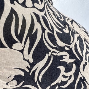 Pu&ograve; includere: Primo piano di un tessuto fantasia nero e beige. Il design presenta un motivo ripetuto di forme astratte a forma di foglia in beige su sfondo nero. Il tessuto sembra leggero e possibilmente seta o un materiale simile.