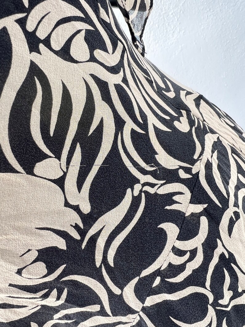Pu&ograve; includere: Primo piano di un tessuto fantasia nero e beige. Il design presenta un motivo ripetuto di forme astratte a forma di foglia in beige su sfondo nero. Il tessuto sembra leggero e possibilmente seta o un materiale simile.