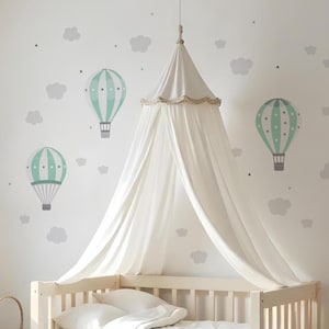 Può includere: Una scena di cameretta con una culla bianca, un baldacchino e adesivi murali. Il baldacchino è drappeggiato sulla culla e la parete è decorata con nuvole grigie, stelle e mongolfiere turchesi e bianche. Il tema è una cameretta fantasiosa.