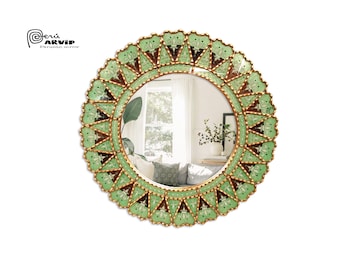 Op de afbeelding: Een ronde spiegel met een decoratief groen en gouden frame. Het frame is gemaakt van kleine, ingewikkelde stukjes glas en metaal. De spiegel reflecteert een kamer met een raam en meubels.