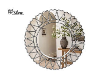 Op de afbeelding: Een ronde zilveren spiegel met een decoratief parelrand. De lijst is van metaal en heeft een geometrisch ontwerp. De spiegel reflecteert een kamer met een vaas met planten op een tafel.