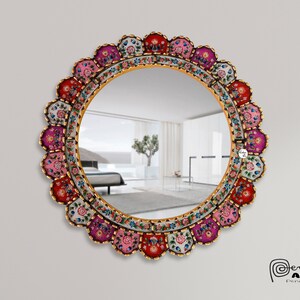 May include: A round mirror with a colorful, floral, and geometric design. The frame is made of wood and painted in shades of pink, red, white, and gold. The mirror is reflecting a room with a white couch and a plant.