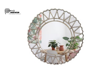 Op de afbeelding: Een ronde, goudkleurige spiegel met een decoratief, geometrisch frame. Het frame is gemaakt van metaal en heeft een zonnestralenontwerp. De spiegel reflecteert een kamer met planten en een tafel.