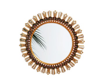 Op de afbeelding: Een ronde gouden spiegel met een decoratief zonnestralenframe. Het frame is versierd met kleine roze en zwarte bloemen.