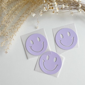 Könnte beinhalten: Drei violette Smiley-Aufkleber auf weißen Papierbögen. Die Aufkleber sind rund und haben ein einfaches Smiley-Gesichtsdesign.